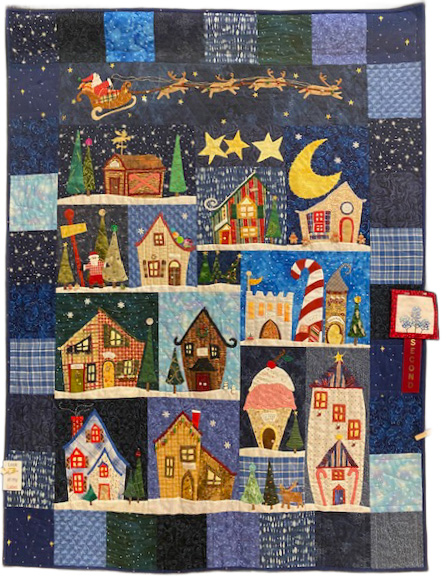A colorful quilt featuring a whimsical winter village scene with various houses, trees, and a sleigh in the night sky.