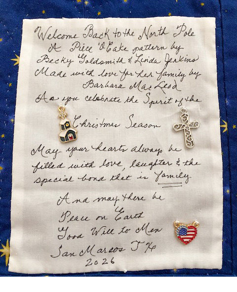 A fabric piece featuring a handwritten holiday greeting with decorative charms, including a church, cross, and heart with an American flag. The message welcomes readers to the Christmas season and emphasizes love and family.